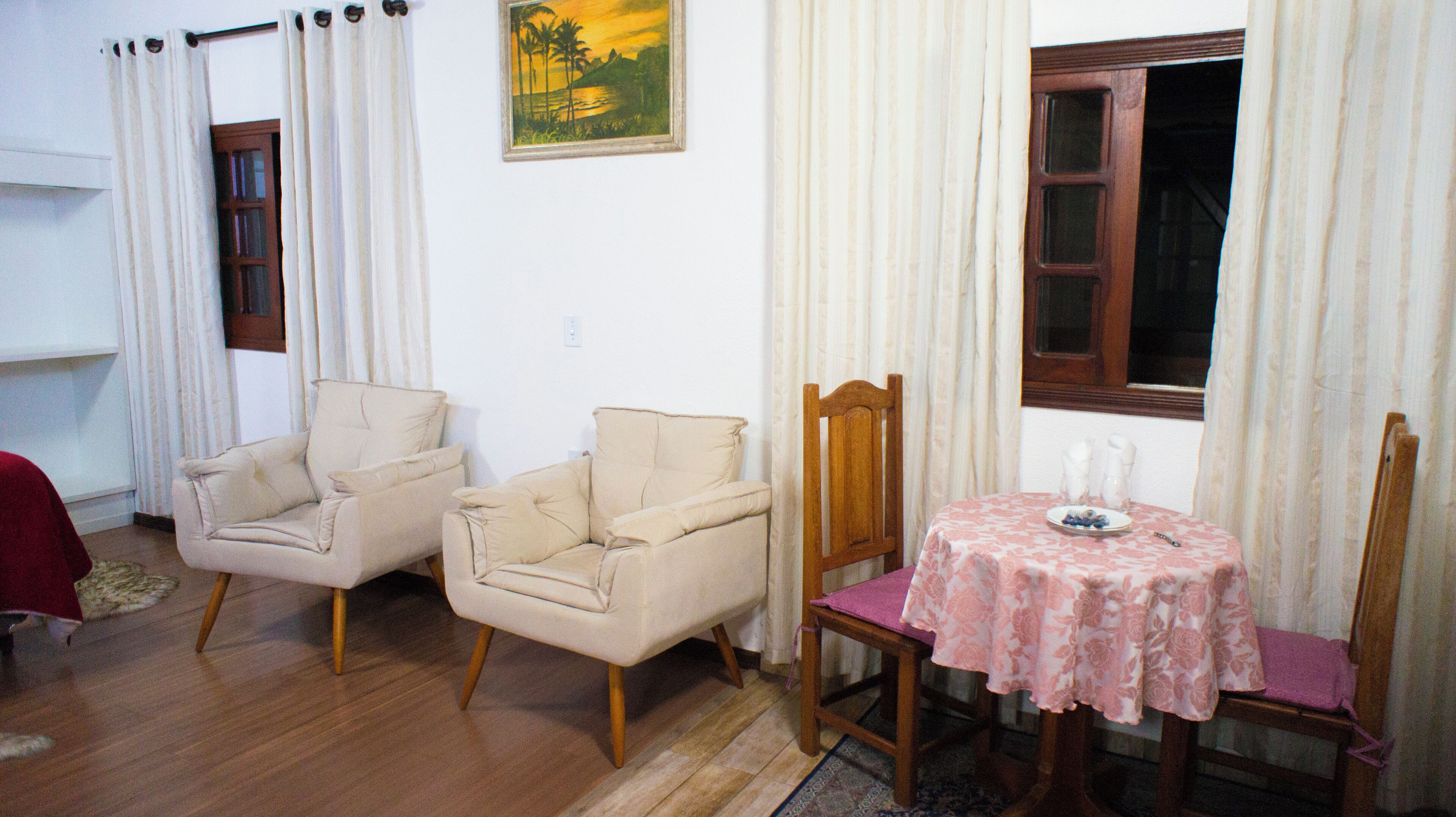 A room with two beige armchairs, a wooden chair, a small table with a pink floral tablecloth, and a window with curtains.