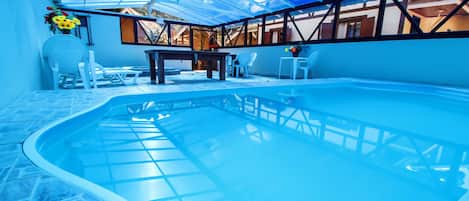 An indoor swimming pool with a glass ceiling, surrounded by white chairs and a table.