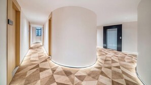 Ein moderner Hotelgang mit geometrisch gemustertem Boden, einer zentralen Säule und einer Tür zum Aufzug.