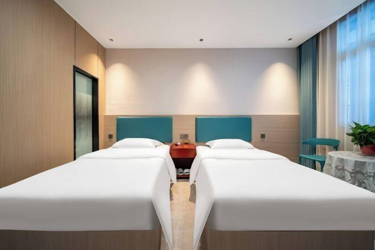 Habitación de hotel con dos camas, una mesita con una planta y vistas al exterior a través de amplias ventanas.