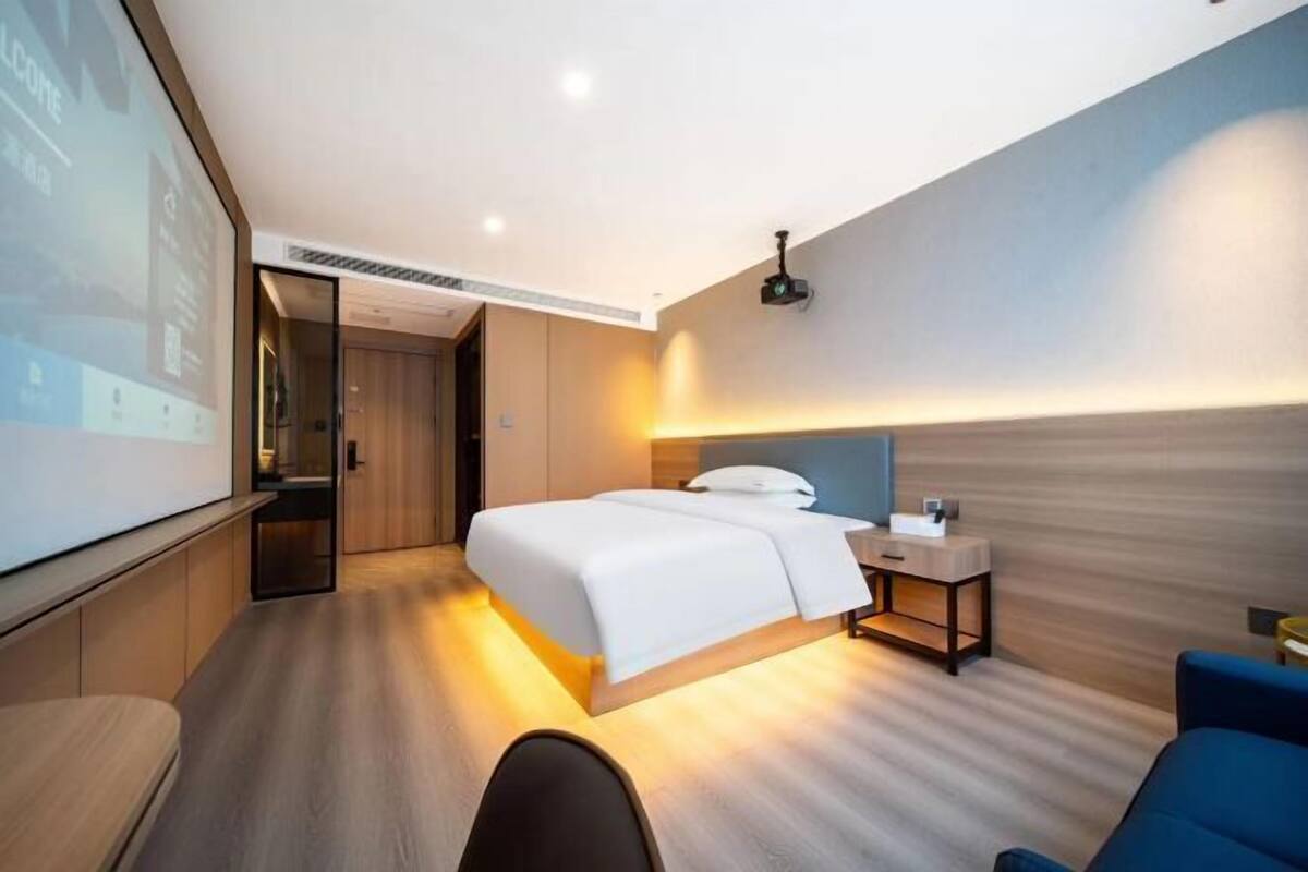 Habitación de hotel moderna con una cama grande, mesita de noche y una pantalla de proyector mostrando un mensaje de bienvenida.