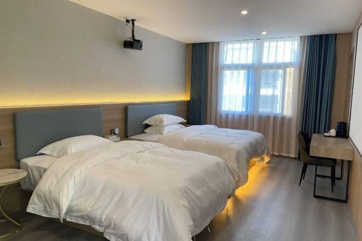 Habitación de hotel con dos camas, una mesita, un proyector y una ventana con cortinas.