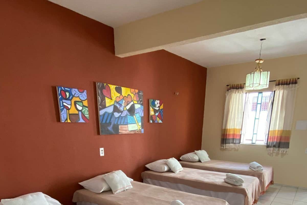 Habitación de hotel con cuatro camas individuales, cada una con cubrecama rosa y almohadas blancas. Hay cuadros coloridos en la pared, una ventana con cortinas y un candelabro.