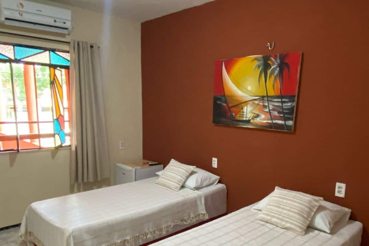 Una habitación con dos camas individuales, un cuadro de un atardecer, una ventana con cortinas y un equipo de aire acondicionado.