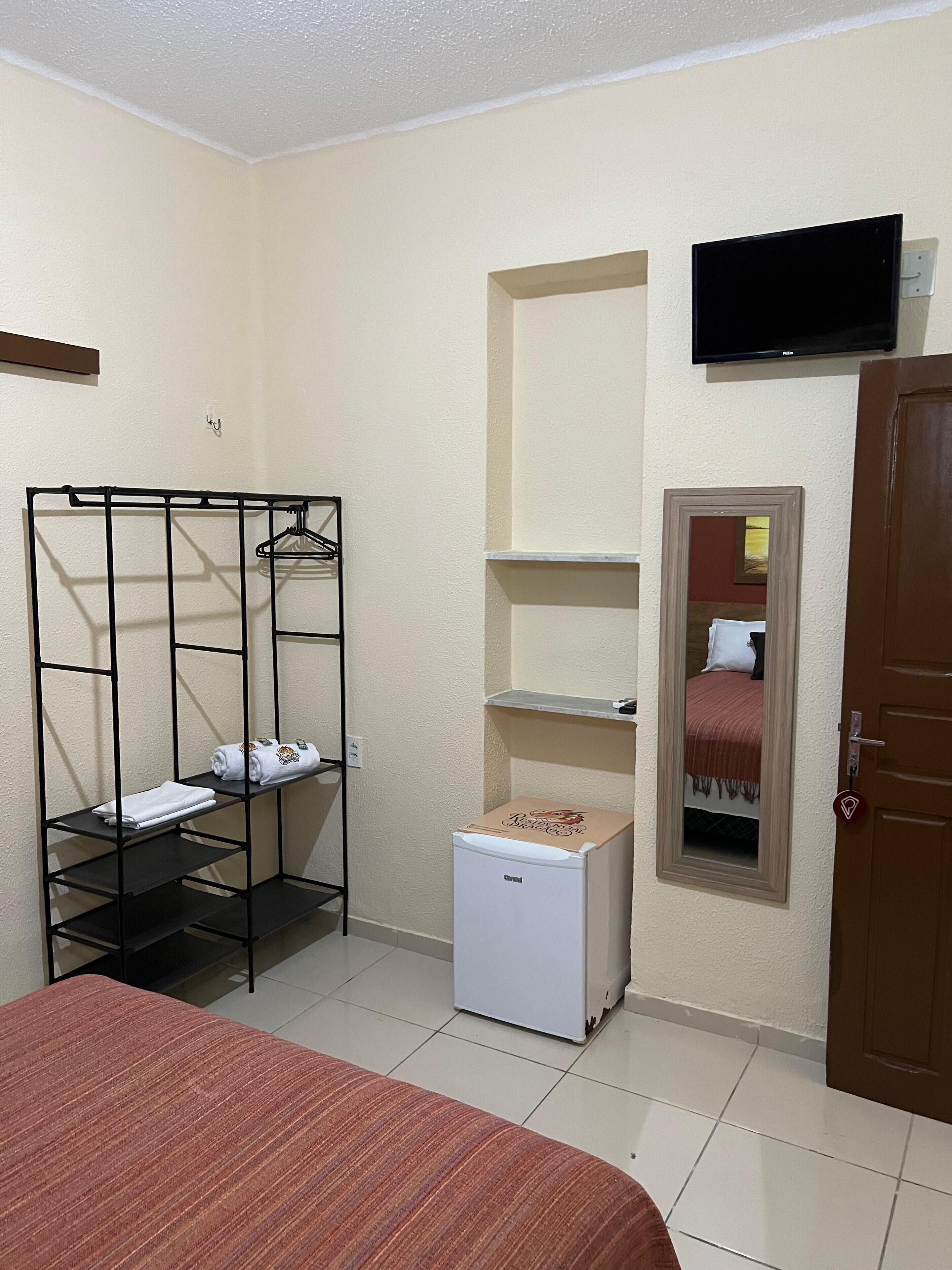 A compact room with a bed, a small refrigerator, a TV mounted on the wall, and a wardrobe with shelves.