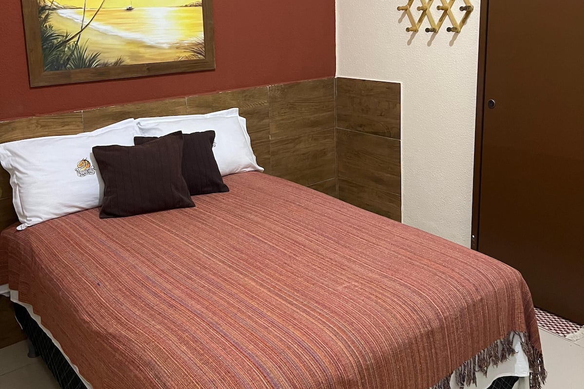Una habitación de hotel con una cama, un cuadro de una puesta de sol, una ventana pequeña y una puerta.