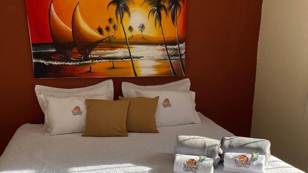 Una cama bien hecha con colcha blanca, dos almohadas y una manta color arena. En la pared detrás de la cama, un cuadro de un atardecer con palmeras.