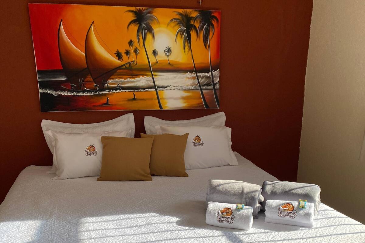 Una cama bien hecha con colcha blanca, dos almohadas y una manta color arena. En la pared detrás de la cama, un cuadro de un atardecer con palmeras.
