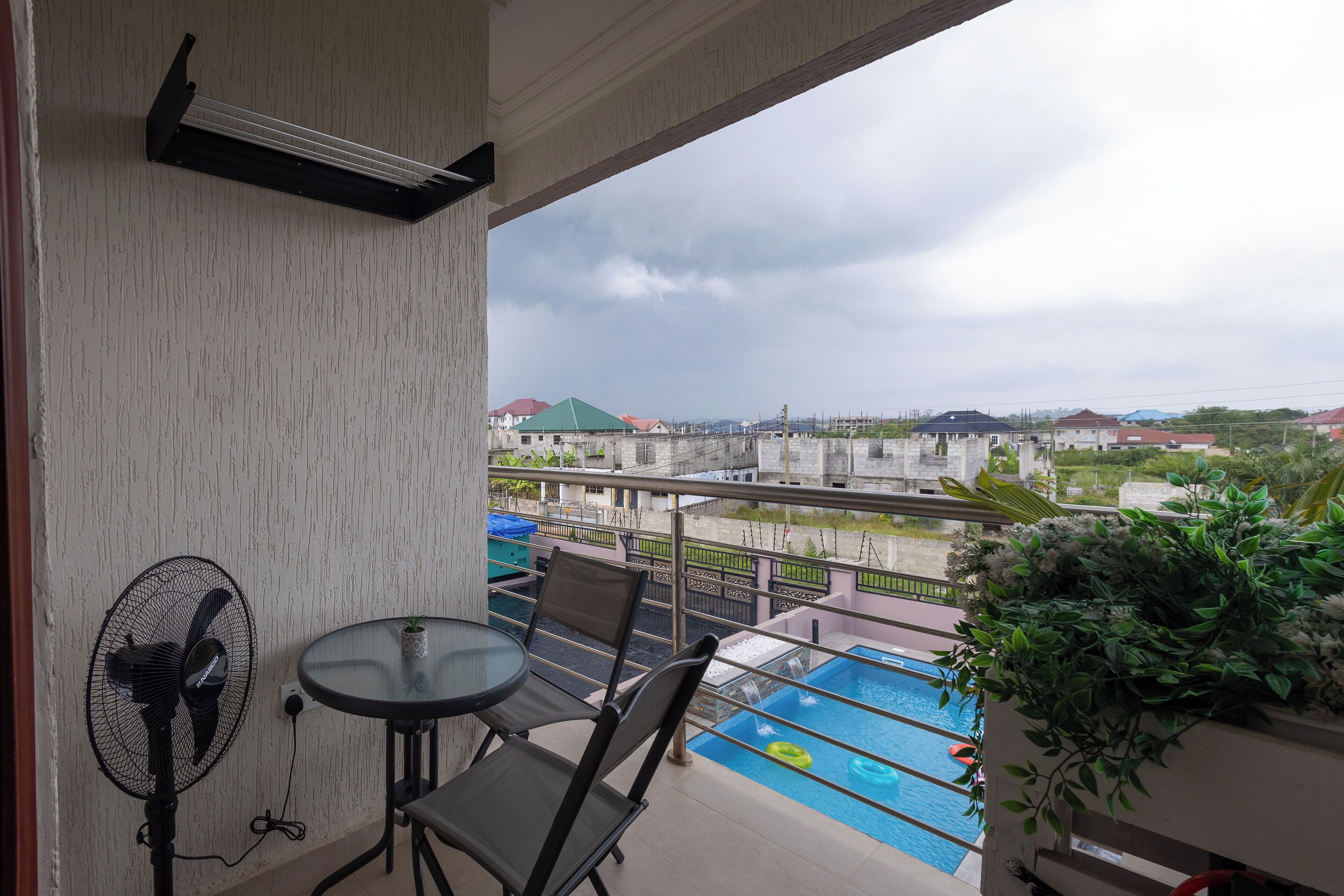 A balcony with a pool, a table, chairs, a fan, and a view of surrounding buildings.