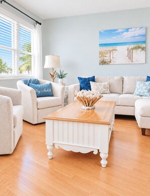 A living room with a white coffee table, beige sofas, and a beach-themed painting. - Family-Friendly Beach Home, 3BR 3BA, Pool, Game Room & Mini Golf (Fernandina Beach)