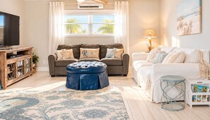 A living room with a sofa, ottoman, and a television on a wooden stand. - Family-Friendly Beach Home, 3BR 3BA, Pool, Game Room & Mini Golf (Fernandina Beach)