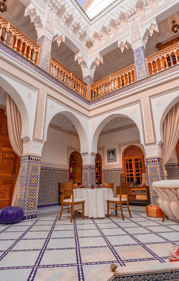 A courtyard with a white tiled floor, wooden furniture, and ornate arches.