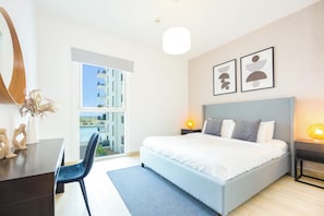 A modern bedroom with a large bed, a desk, and a chair. There is a window with a view of buildings and a balcony.