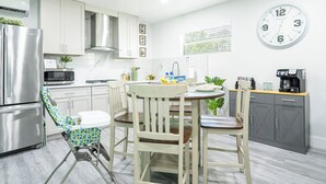 A modern kitchen with white cabinets, a stainless steel refrigerator, a microwave, and a coffee machine. There is a dining table with chairs and a small folding chair. - Cozy & Stylish Apartment for 4 just 5 Minutes from Downtown (Mount Dora)