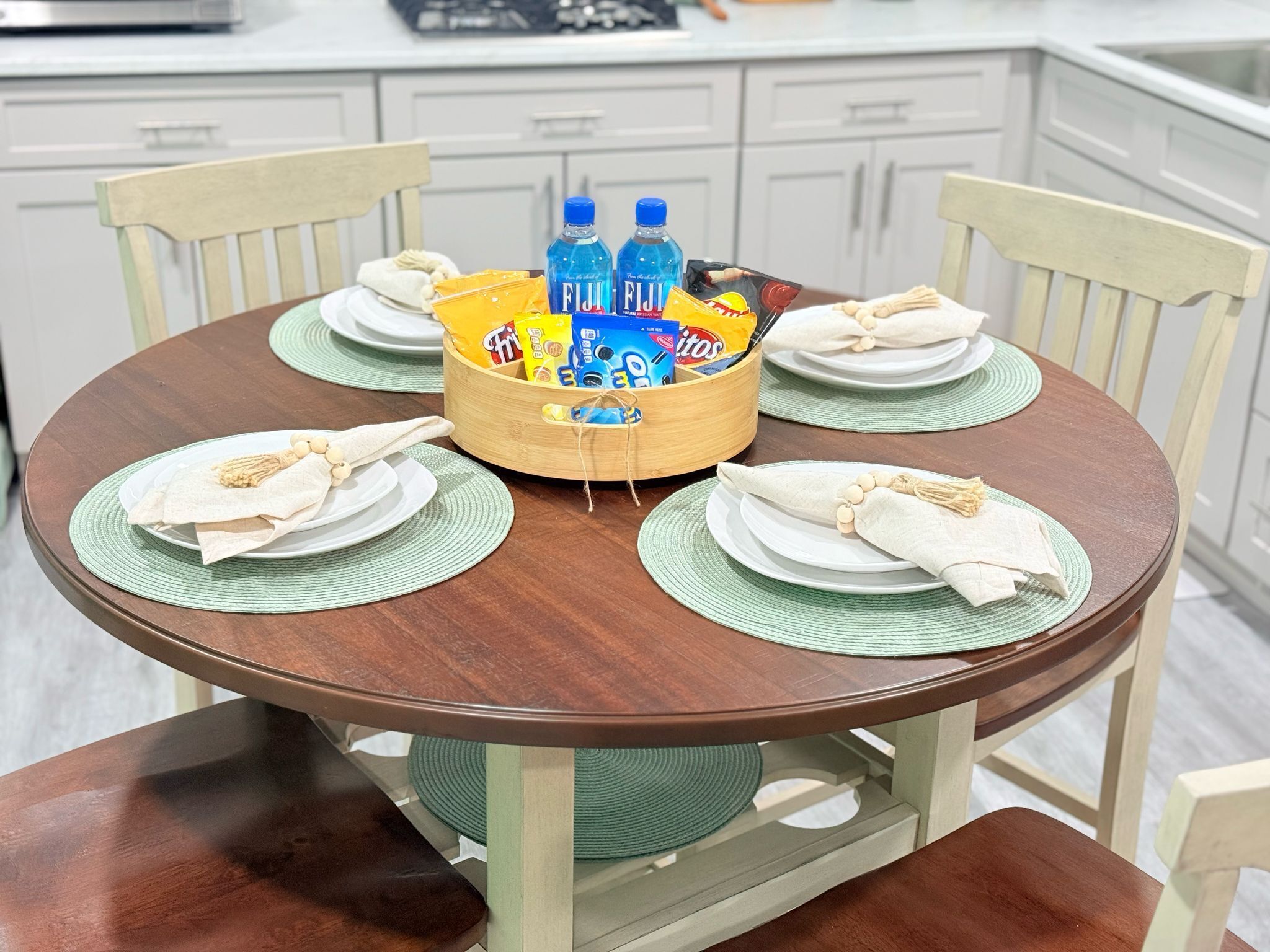 Une table de salle à manger ronde en bois, agrémentée de sets de table verts, d’assiettes blanches et de serviettes. Un panier en bois placé au centre contient des bouteilles d’eau et des en-cas.