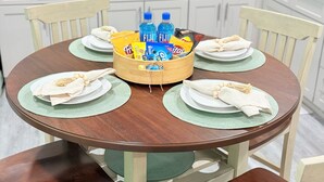 A round wooden dining table with green placemats, white plates, and napkins. A wooden basket in the center holds bottled water and snacks. - Cozy & Stylish Apartment for 4 just 5 Minutes from Downtown (Mount Dora)