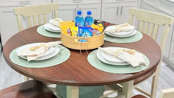 Une table de salle à manger ronde en bois, agrémentée de sets de table verts, d’assiettes blanches et de serviettes. Un panier en bois placé au centre contient des bouteilles d’eau et des en-cas.