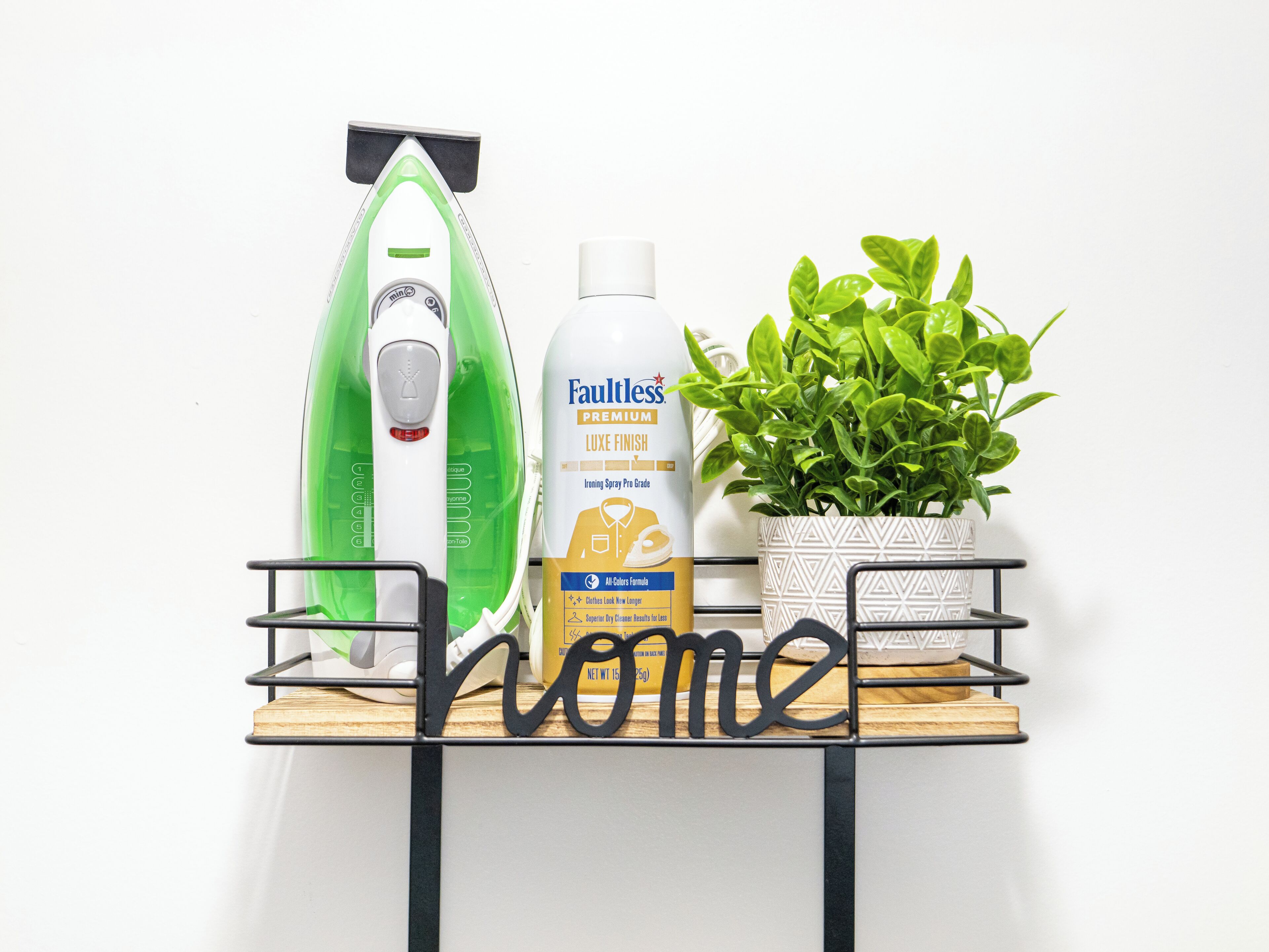 A shelf with an iron, a bottle of Faultless Premium Luxe Finish Ironing Spray, and a potted plant.