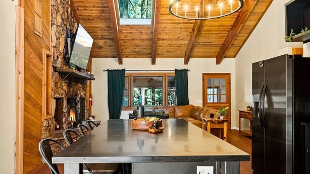 A rustic kitchen with a stone fireplace, wooden beams, a large island table, and a modern refrigerator.