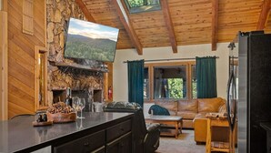 A cozy cabin interior with a stone fireplace, wooden ceiling, a television mounted above it, a sofa, and a dining area with a wooden table.