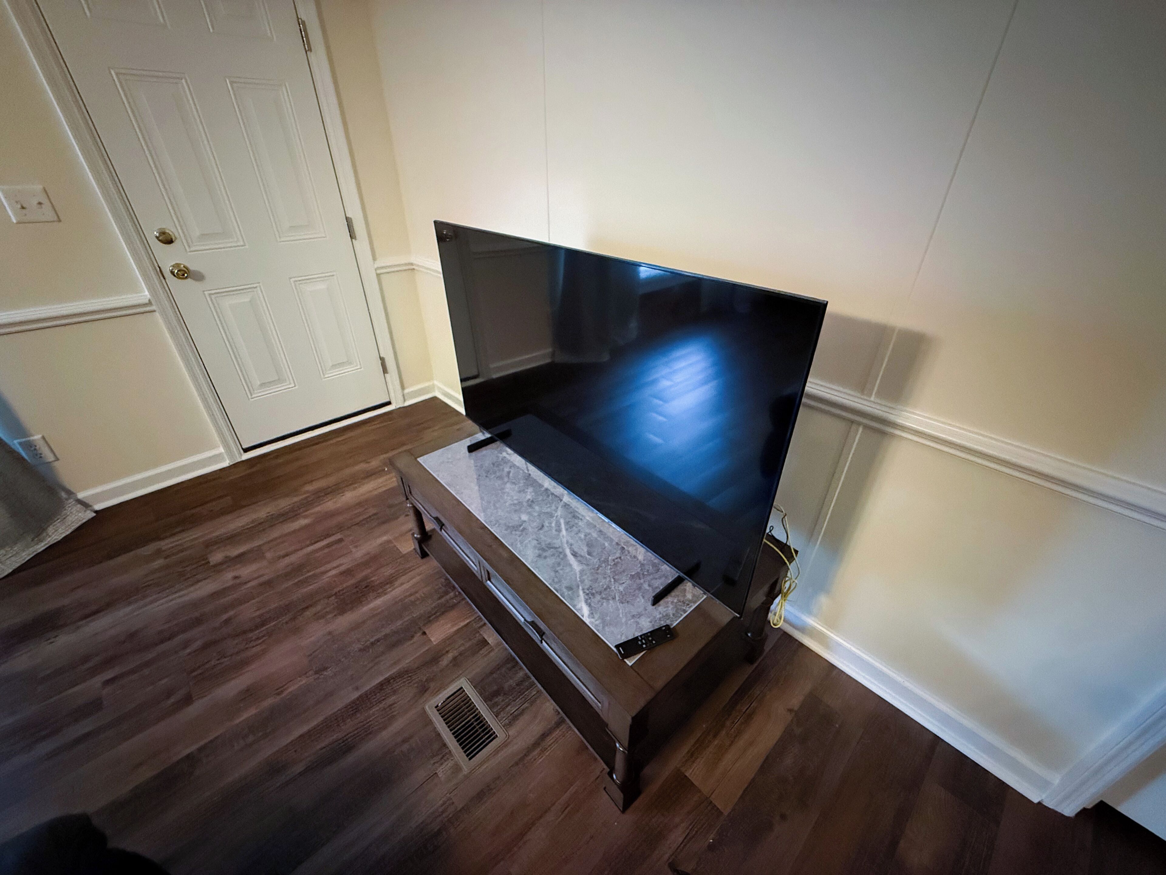 A room with a large flat-screen TV on a stand, wooden flooring, a white door, and a vent on the floor.
