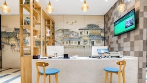 Un espace d’accueil comprenant un comptoir, deux tabourets en bois, un écran numérique et une fresque représentant des bâtiments traditionnels.