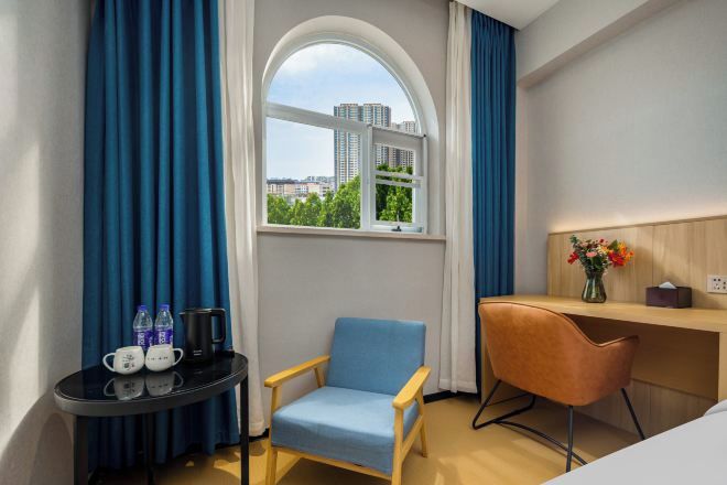 Quarto de hotel com uma grande janela em arco, uma escrivaninha de madeira, uma cadeira azul, uma pequena mesa com chaleira e bules, e um vaso com flores.