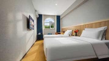 A hotel room with two beds, a TV mounted on the wall, a window with curtains, and a view of greenery outside.