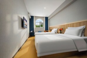 A hotel room with two beds, a TV mounted on the wall, a window with curtains, and a view of greenery outside.