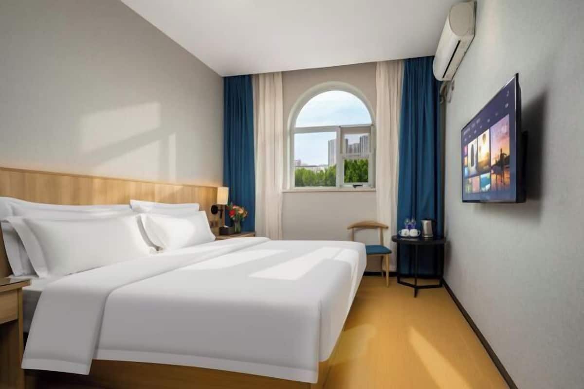 Une chambre d’hôtel avec un grand lit, une petite table avec une chaise, une fenêtre avec des rideaux et un téléviseur fixé au mur.