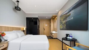A hotel room with a bed, bedside table, a small table with a kettle and bottles, a chair, a large framed picture on the wall, and a door leading to another room.