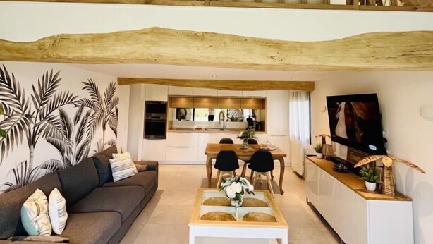 Een ruime loft met zichtbare houten balken, een gezellige zithoek met een bank en voetenbankje, een eettafel, en een keuken met een televisie.