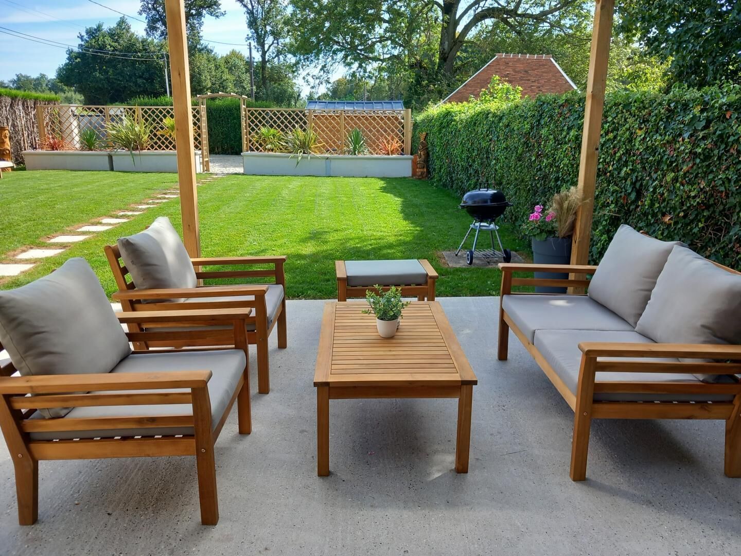 A patio with wooden furniture, a table with a plant, and a barbecue grill in the background.
