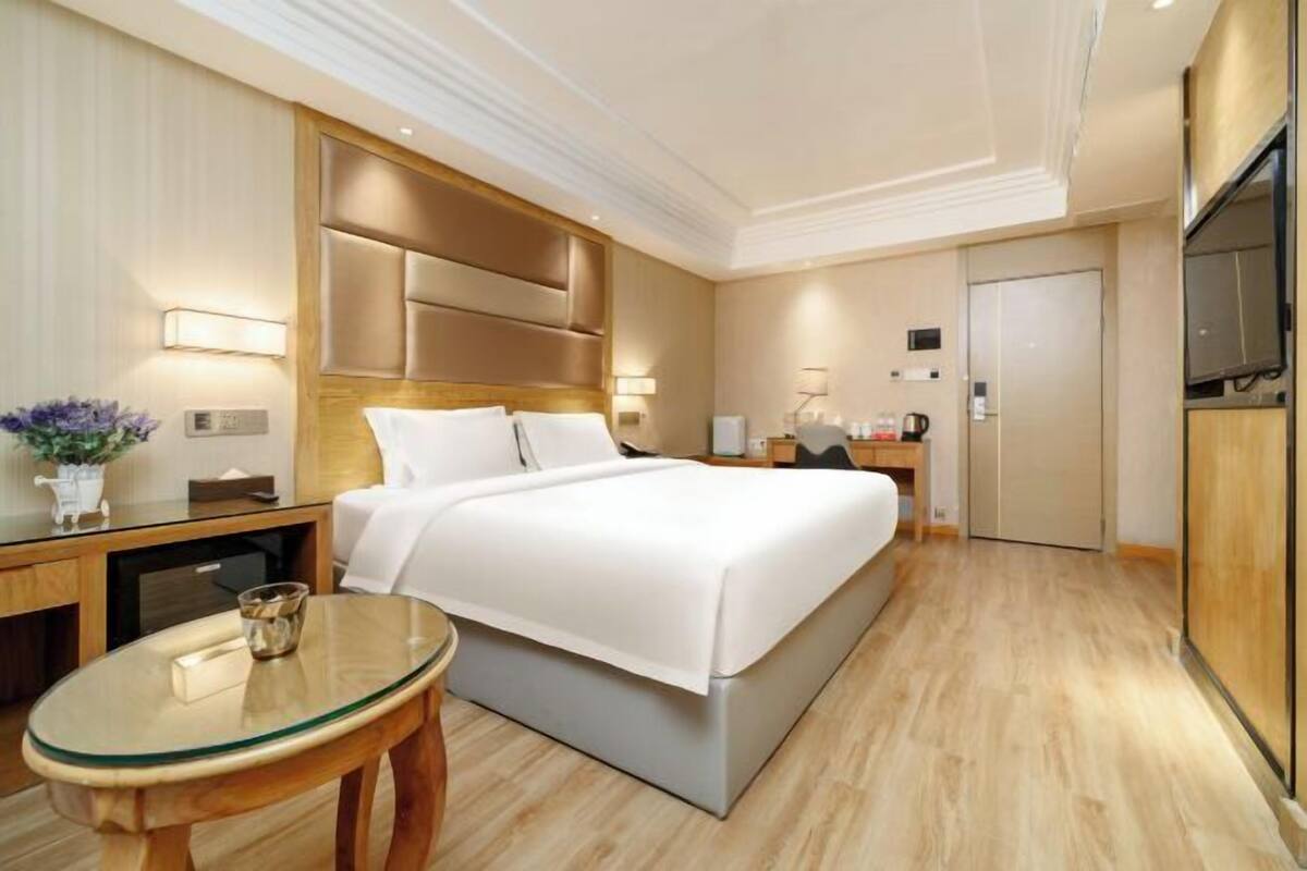 A modern hotel room with a large bed, a bedside table, a glass table, and a wardrobe.
