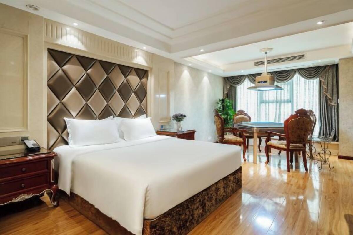 A hotel room with a large bed, a dining table with chairs, a small side table, and a large window with curtains.