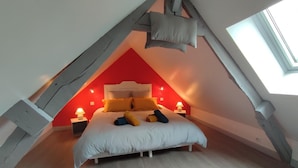 A bedroom with a sloped ceiling, a bed with pillows, two bedside lamps, and a window. - Gîtes de France® - Le Petit Marin (BRUNVILLE)