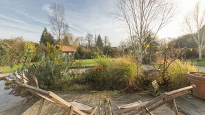 A wooden deck with two wooden chairs, a pond, and a garden with various plants and trees. - Gîtes de France® - Le Petit Marin (BRUNVILLE)