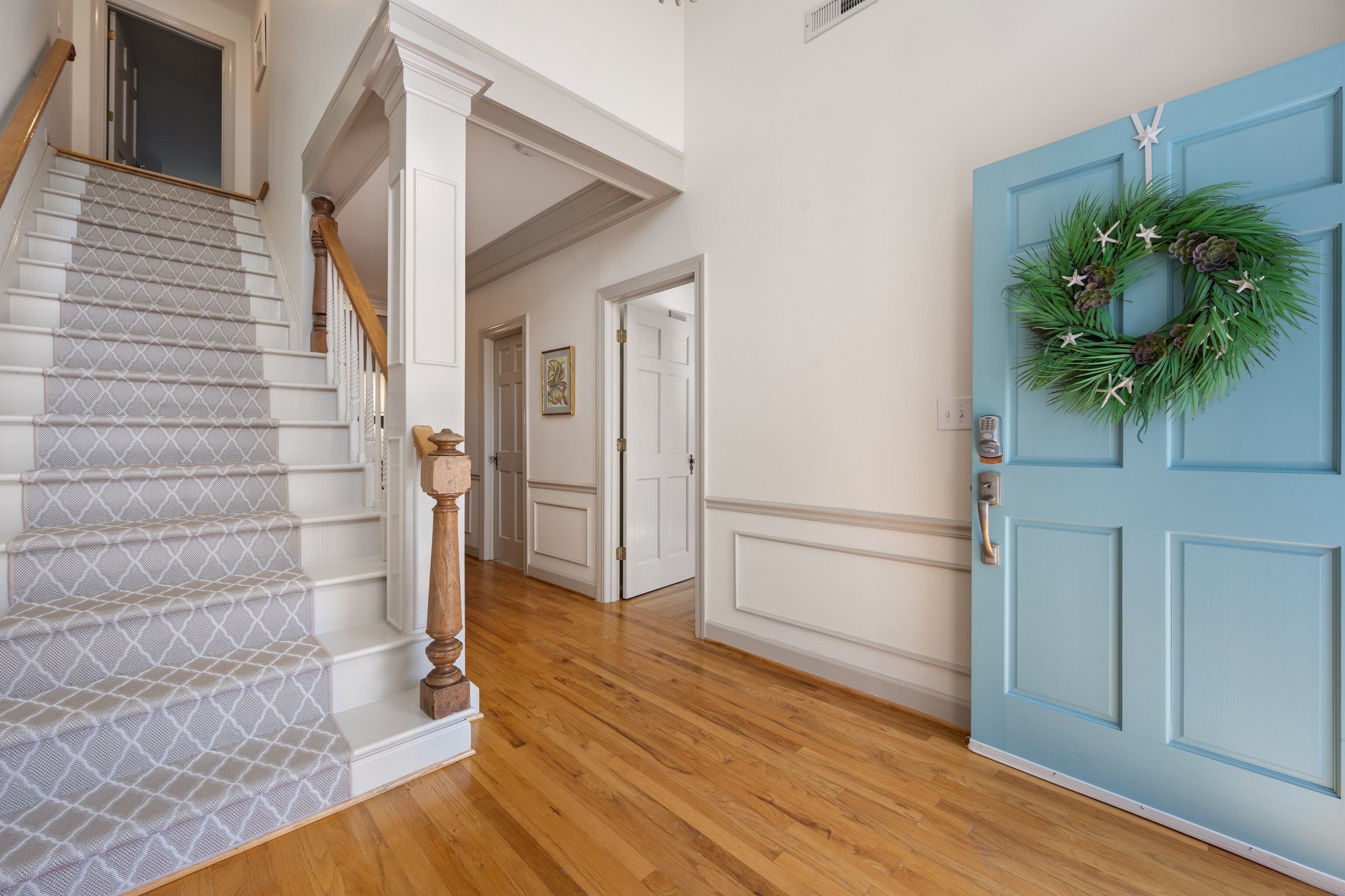 Un escalier recouvert d’une moquette à motifs, une porte bleue ornée d’une guirlande, et un couloir aux murs blancs et au plancher en bois.