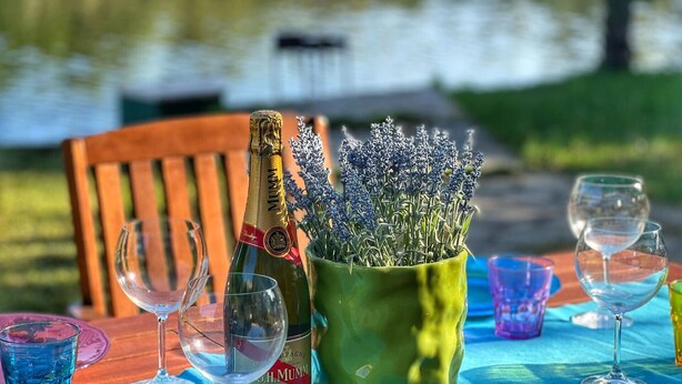 A table with a bottle of champagne, a green vase with lavender, and several wine glasses on a blue tablecloth by a body of water.