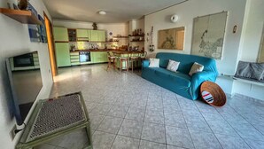 A living room with a blue sofa, a dining table with chairs, a kitchen in the background, and a TV on the left.