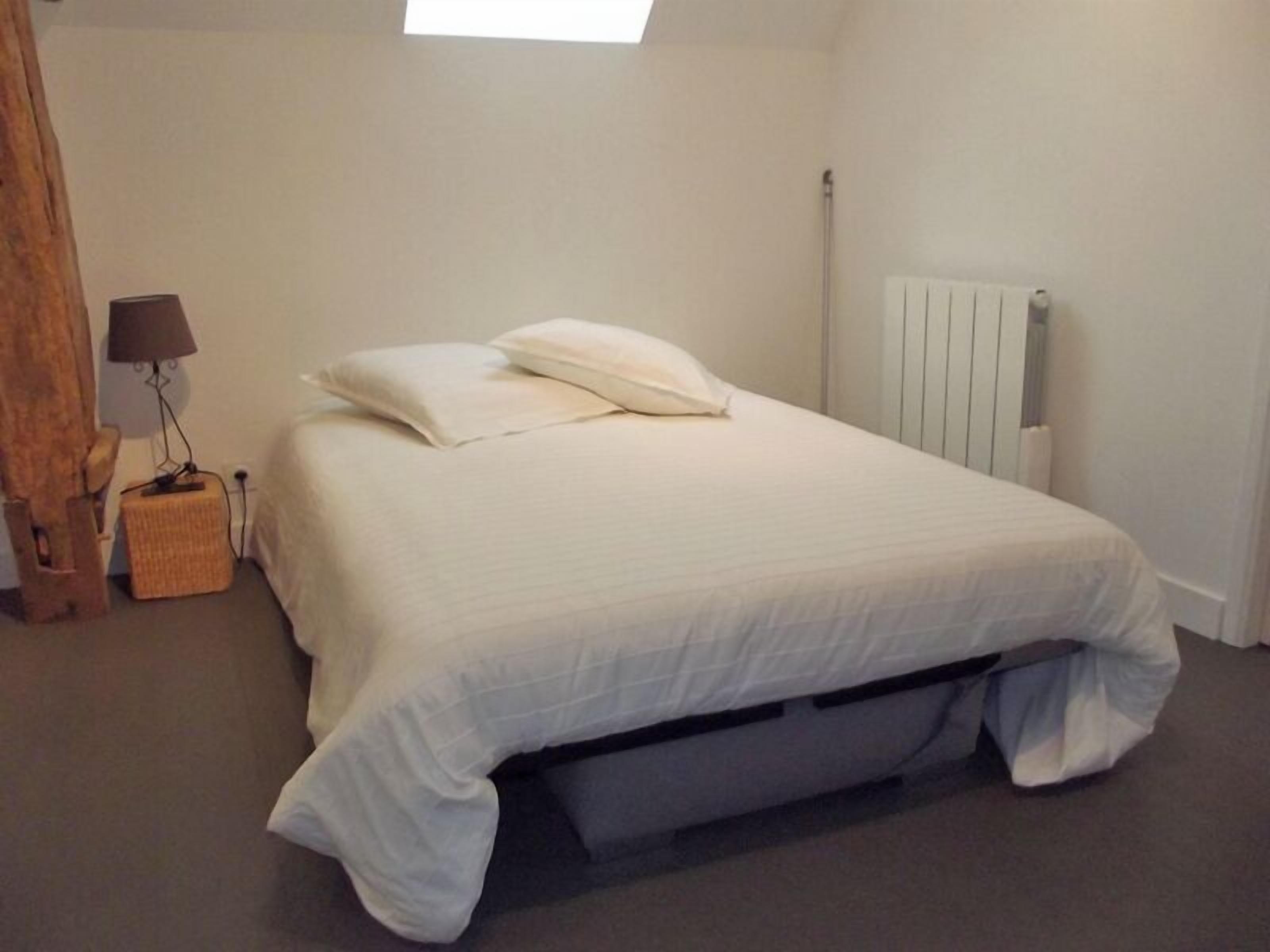 Un dormitorio con una cama, un radiador, una lámpara y un taburete de madera.
