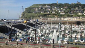 Un port de plaisance où sont amarrés de nombreux bateaux, une promenade piétonne et une colline avec des bâtiments en arrière-plan.