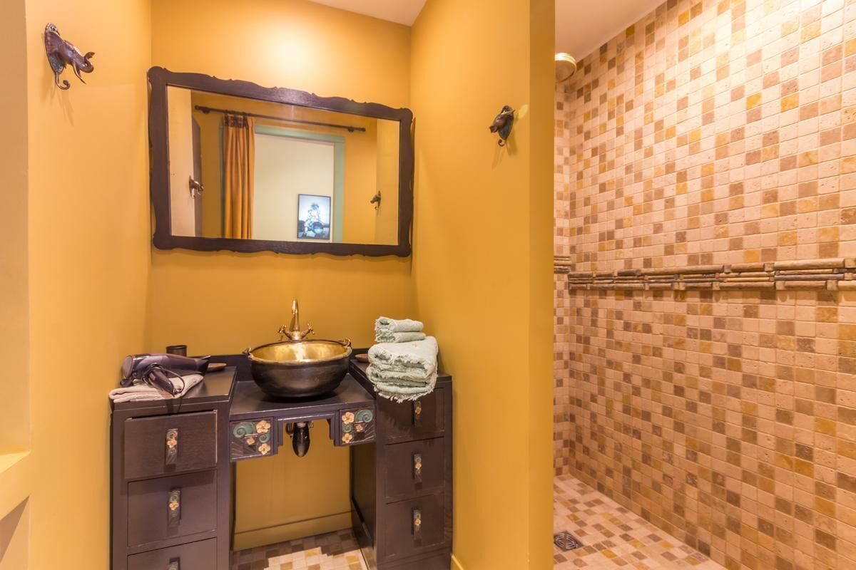 Une salle de bain avec un mur jaune, un lavabo avec un receveur, un miroir et une zone de douche carrelée.