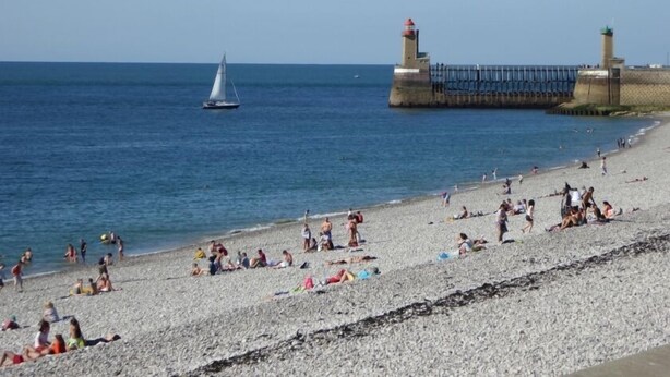 Une plage où des gens se prélassent au soleil et où l’on aperçoit un voilier sur l’eau.