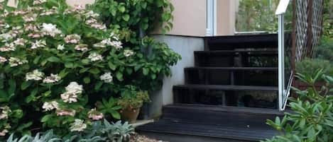 A modern building with a beige exterior, a staircase leading to a door, and a garden area with shrubs and pebbles.