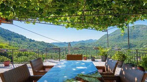 A terrace with wicker furniture, a table, and potted plants, overlooking a mountainous landscape.