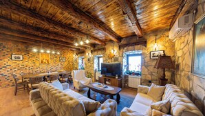 A cozy living room with a stone wall, wooden ceiling beams, and a fireplace. There is a television, a dining table with chairs, and a sofa.