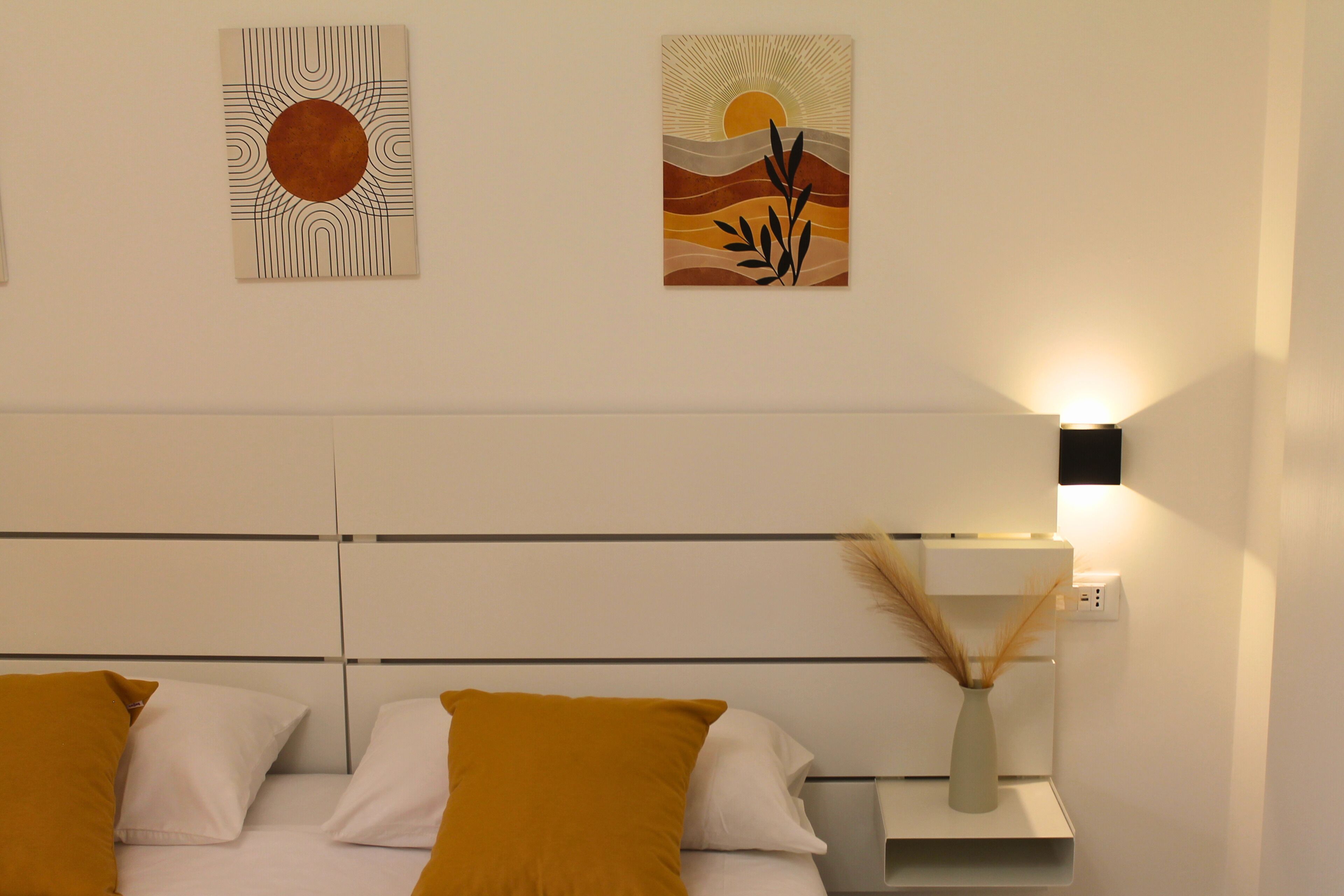 Quarto de hotel com uma cama, mesa de cabeceira e duas obras de arte fixadas na parede representando cenas abstratas de sol e paisagem.