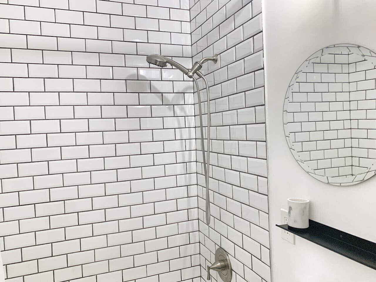 A bathroom with white subway tile walls, a shower with a handheld showerhead, a toilet, and a black shelf with a white cup.