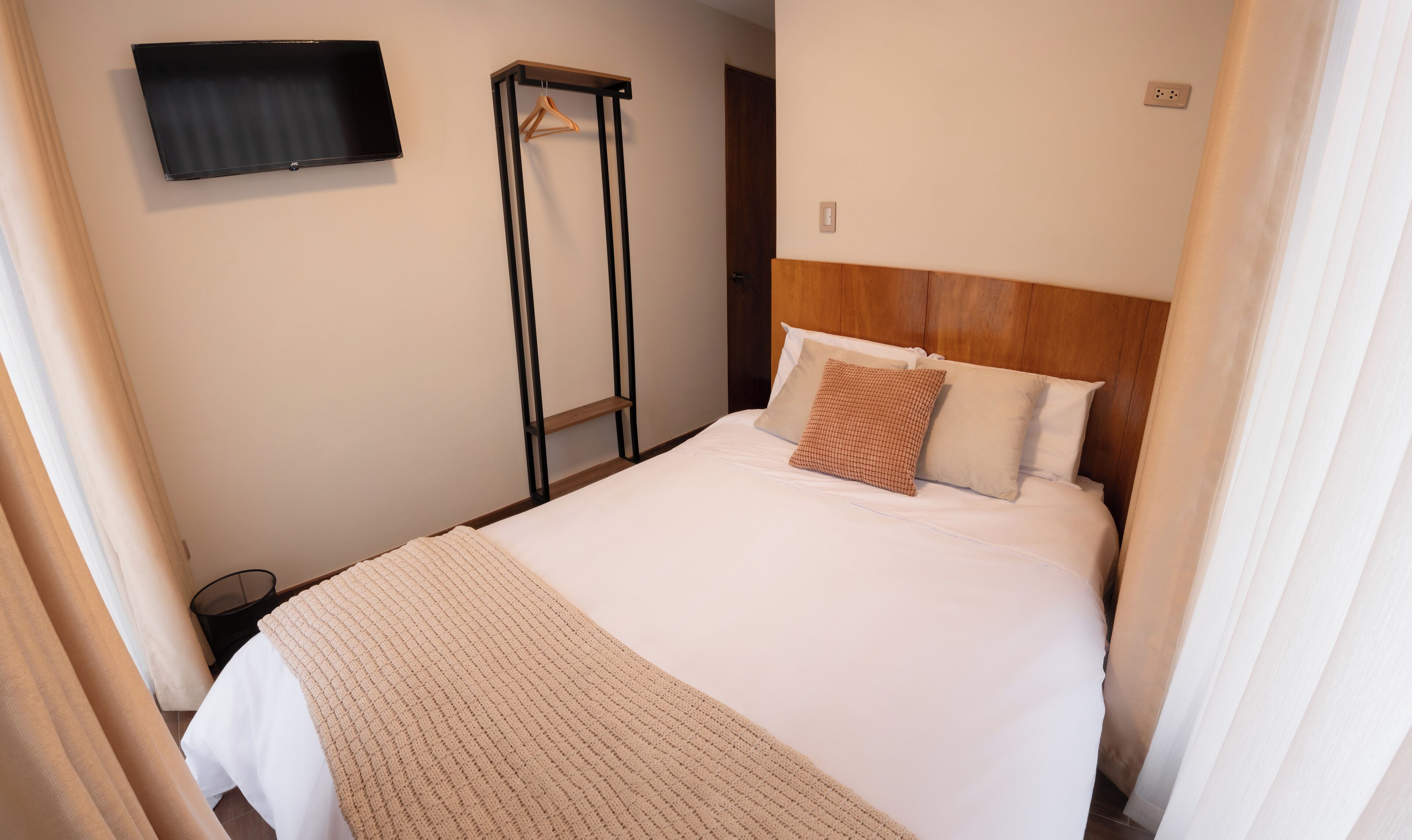 A neatly made bed with a white comforter and two pillows, a wooden headboard, a television mounted on the wall, and a hanging rack.